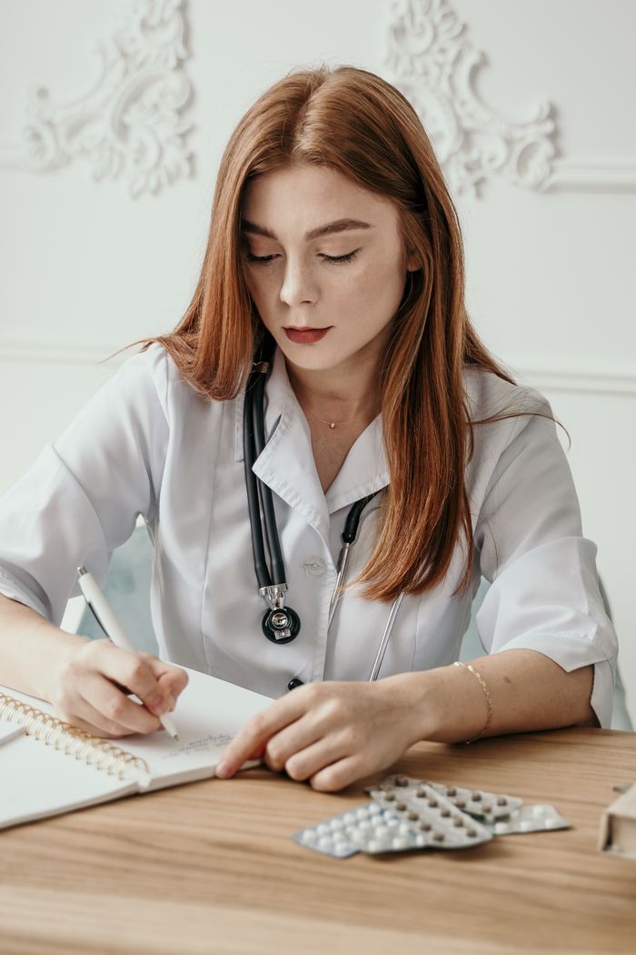 About Us Young female doctor writing notes at her desk with stethoscope and medication in view.