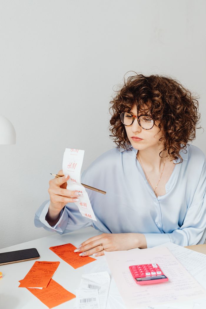 Offerings Curly-haired woman examining receipts with a calculator at her desk.